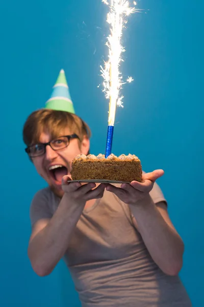 Estudante cara alegre louco em óculos cumprimentando com um chapéu de papel segurando um bolo com fogos de artifício em chamas em suas mãos. Conceito de aniversário feriado — Fotografia de Stock