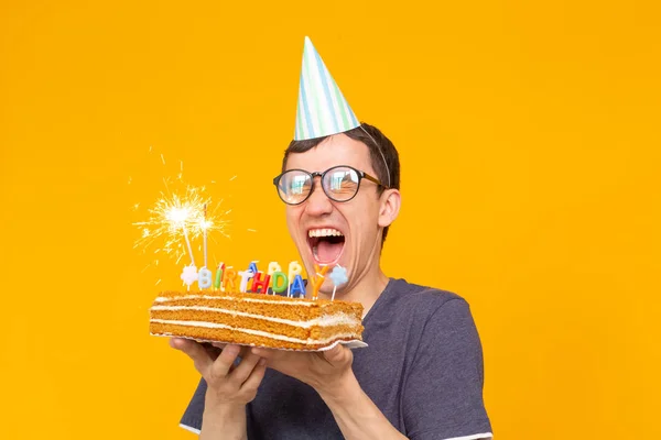 Divertente ragazzo positivo in occhiali tiene tra le mani una torta fatta in casa con l'iscrizione buon compleanno in posa su uno sfondo giallo. Concetto di vacanze e anniversari . — Foto Stock