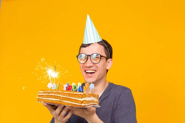 Pazzo allegro giovane ragazzo asiatico con gli occhiali che tiene una candela accesa tra le mani e una torta fatta in casa di congratulazioni su uno sfondo giallo. Concetto di compleanno e anniversario . — Foto Stock