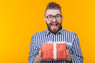 Şaşkın genç hipster adam bıyık ve sakal sürpriz bir sarı arka planda hediyeler ile kırmızı bir kutu ayıklar, yakın yukarı. Tatil için hediyeler ve sürprizler kavramı