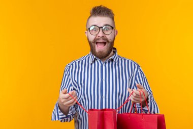 Gözlük ve sakal ile sürpriz neşeli genç yakışıklı adam sarı bir arka planda ayakta, alışveriş için paketler içine bakıyor. Başarılı satın alma kavramı. Boşaltmak.