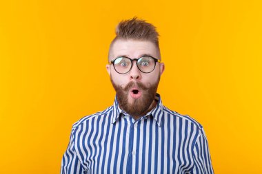 Bıyıklı ve sakallı genç adam, sarı bir arka plan karşısında sürpriz bir şekilde ağzını yuvarladı. Sürpriz ve belirsizlik kavramı.