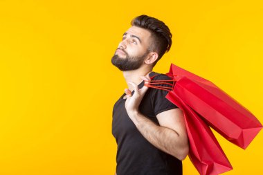 Sarı bir arka plan üzerinde poz alışveriş çantaları tutan üzgün bir genç sakallı şık hipster adam Yan görünümü. Gereksiz satın alma kavramı.