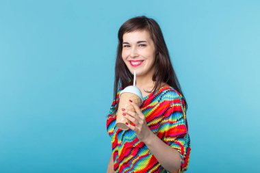 Modaya uygun parlak giysi aksesuarları giyen ve makyaj güzel hipster kız Komik portre. O kopya alanı ile mavi arka plan üzerinde smoothie içiyor