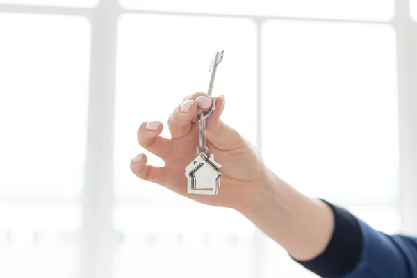 Mortgage concept. Female hand holding key with house shaped keychain. Modern light interior. Real estate, moving home or renting property. — Stock Photo, Image