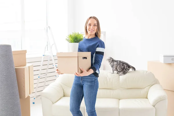 Очаровательная молодая незамужняя женщина держит коробку с вещами во время переезда, стоя в новой квартире рядом со своим седоухим шотландским котом. Концепция переезда и новоселья . — стоковое фото