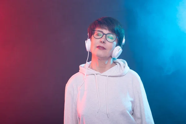 Beautiful middle-aged woman in a white sweater and glasses is listening to music while posing on a red-blue background in a studio. The concept of a favorite hobby. Advertising space. — Stock Photo, Image