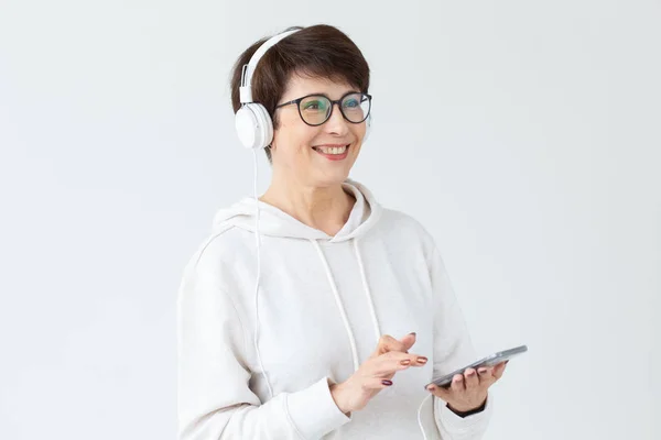 Cute positive middle-aged woman in sweater and glasses is listening to music with wire headphones standing on a white background and holding tablet . Concept of hobbies and subscriptions to favorite — Stock Photo, Image