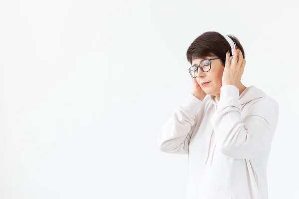 Cute positive middle-aged woman in sweater and glasses is listening to music with wire headphones standing on a white background with copy space. Concept of hobbies and subscriptions to favorite radio — Stock Photo, Image