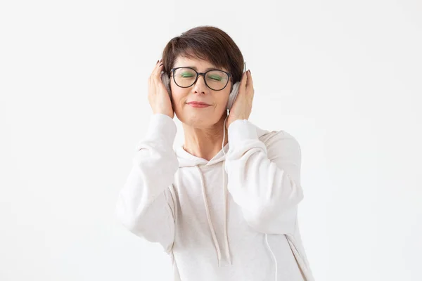 Mujer de mediana edad bastante entusiasta en gafas y un suéter blanco escucha su música favorita con auriculares sobre un fondo blanco con espacio para copiar. Concepto de suscripción de radio y música en línea . — Foto de Stock