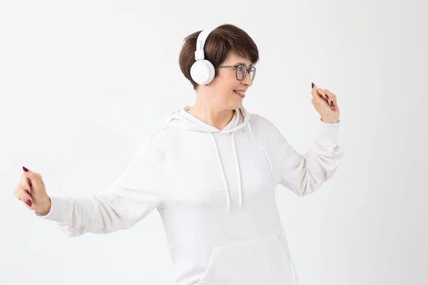 Pretty keen middle-aged woman in glasses and a white sweater listens to her favorite music with headphones on a white background. Online radio and music subscription concept. — Stock Photo, Image