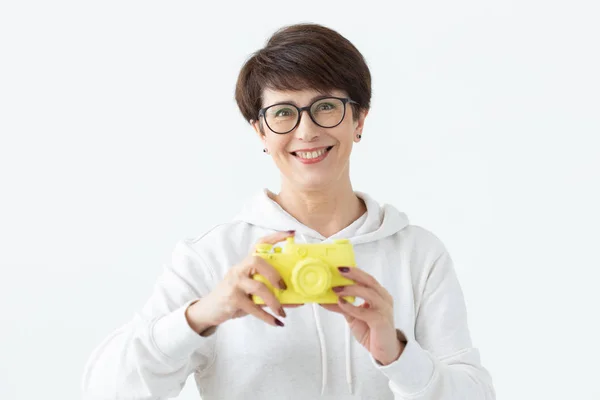 Mujer positiva de mediana edad en gafas sostiene en la mano una cámara amarilla vintage posando sobre un fondo blanco. Concepto de amantes de la foto y aficiones — Foto de Stock