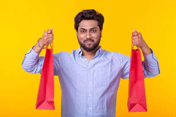 Fiatal csinos indiai férfi a szakállas tart piros bevásárló táskák pózol egy sárga háttér. Szupermarket vásárlási koncepció. — Stock Fotó