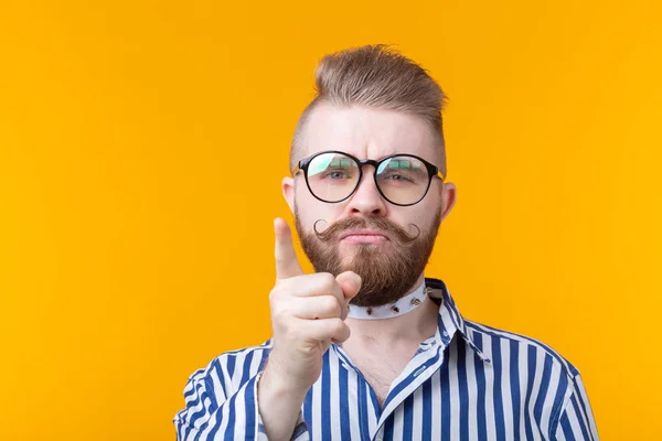 Funny young man with a mustache and a beard and glasses shows on you on a yellow background. Concept of non-standard advertising.