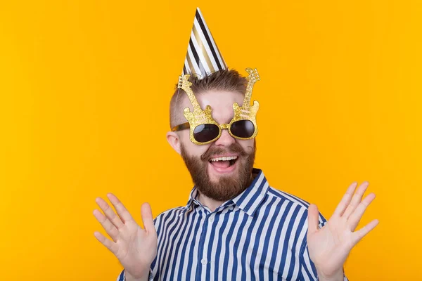 Χαρούμενο νεαρό χίπστερ αρσενικό με χάρτινο καπάκι και γυαλιά γελάει ευτυχισμένα σε κίτρινο φόντο. Η έννοια του Μέγα Κόμματος και των διακοπών. — Φωτογραφία Αρχείου