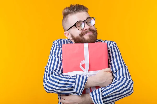 Joyful young hipster guy with a mustache in glasses hugs a red gift box on a yellow background. The concept of the joy of gifts. — Stock Photo, Image