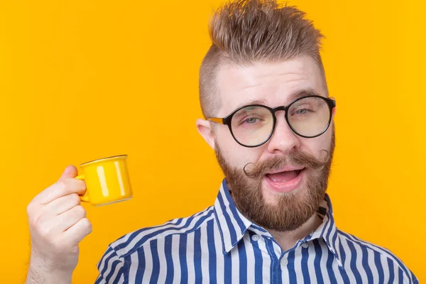 Retrato de un joven guapo divertido y seguro de sí mismo en gafas con una barba con una pequeña taza amarilla sobre un fondo amarillo. Buen concepto del día . — Foto de Stock