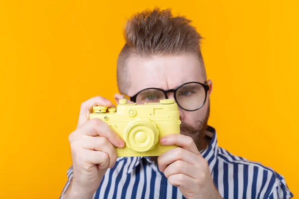 Bıyıklı ve sakallı sevimli genç erkek fotoğrafçı sarı bir arka plan üzerinde sarı bir vintage kamera fotoğraflıyor. Hobi ve profesyonel çalışma kavramı. — Stok fotoğraf