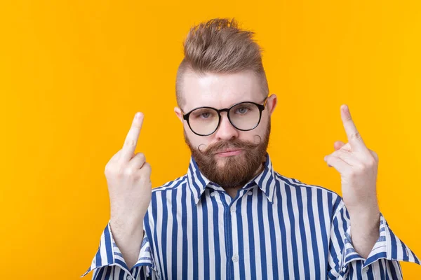 Ein wütender junger Mann mit Brille, Schnurrbart und Bart zeigt den Mittelfinger, der auf gelbem Grund posiert. Konzept der Verleugnung und Verärgerung. — Stockfoto