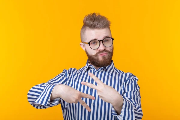 Encantador joven hipster con gafas y barba muestra gesto de victoria posando sobre un fondo amarillo. El concepto de confianza en sí mismo y divertido . — Foto de Stock