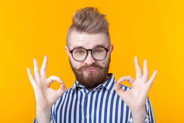 Un joven guapo con bigote y barba con gafas muestra que está bien posando sobre un fondo amarillo. Concepto de suerte y barbería . — Foto de Stock