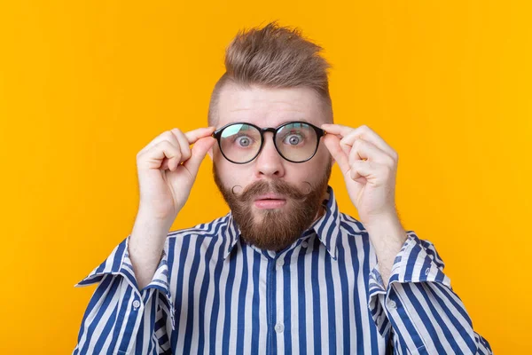 Überraschter junger Hipster mit Schnurrbart und Bart blickt durch eine Brille, die auf gelbem Grund posiert. Konzept der Überraschung. — Stockfoto