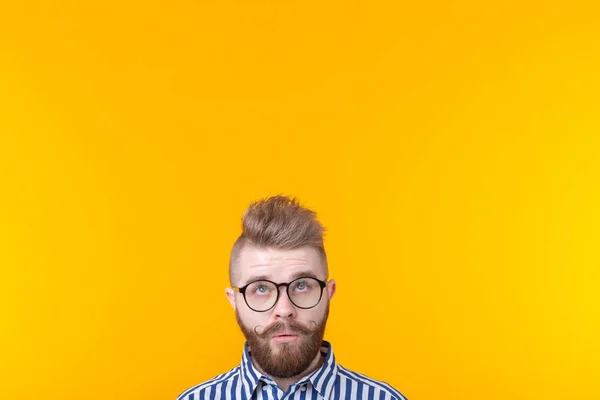 Sorprendido joven hipster hombre con bigote y con gafas posando sobre un fondo amarillo en sorpresa mirando hacia arriba. Concepto de sorpresa y lugar de publicidad y enlaces . — Foto de Stock