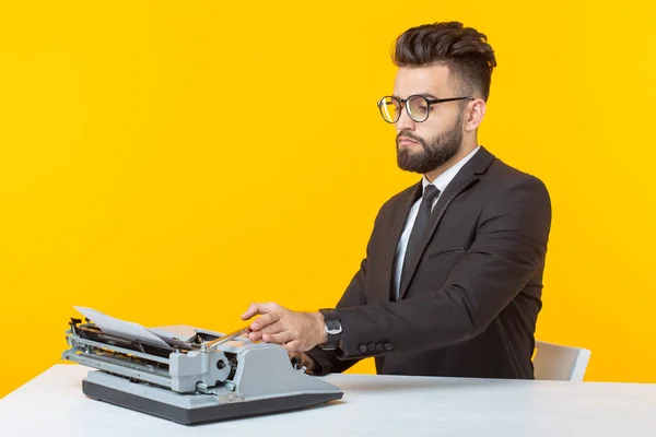 Joven hombre de negocios guapo en ropa formal escribiendo texto en una máquina de escribir posando sobre un fondo amarillo. Lugar para la publicidad. Concepto empresarial . — Foto de Stock