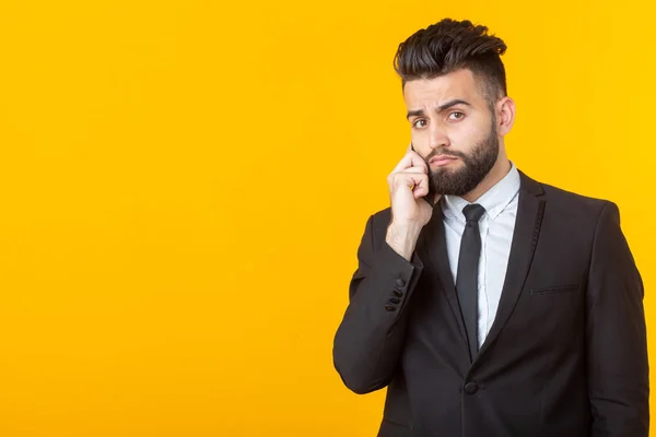 Cute young man with a beard in formal clothes talking on the phone posing on a yellow background with copy space. Concept of an informal meeting and business deal.