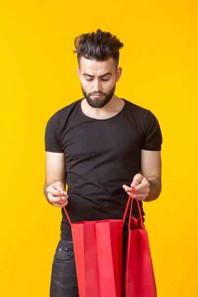 Cute young sad bearded man looks at the purchase in shopping bags on a yellow background with copy space. Concept of rash and waste of money.