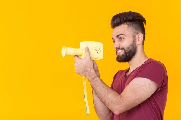 Vista lateral de um jovem hipster bonito com uma barba atira objetos em uma câmera de vídeo vintage em um fundo amarelo com espaço de cópia. Conceito de vídeo — Fotografia de Stock