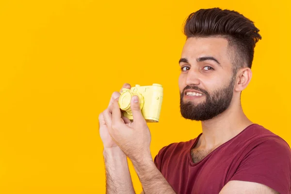 Joven hombre hipster positivo con una barba sosteniendo una cámara vintage amarilla posando sobre un fondo amarillo. Concepto de hobby y fotografía profesional — Foto de Stock