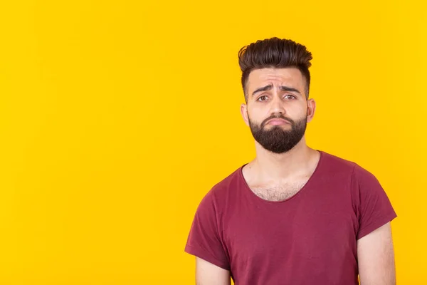 Triste jovem e bonito hipster com uma barba em uma camiseta borgonha posando em um fundo amarelo. Conceito de frustração . — Fotografia de Stock