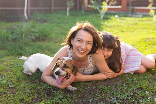 Klein meisje in roze jurk met moeder en Jack Russell Terrier hond leggen op gras. Kijk naar camera en glimlach — Stockfoto