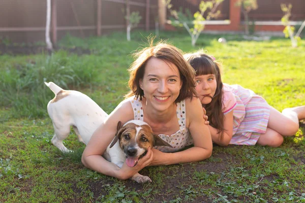 Liten flicka i rosa klänning med mor och Jack Russell Terrier hund om på gräs. Titta på kamera och leende — Stockfoto