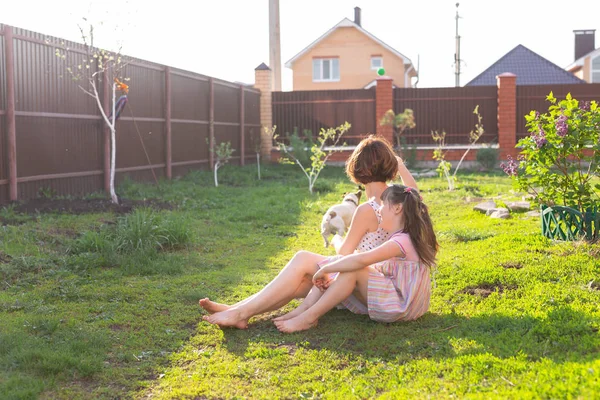 Маленькая девочка в розовом платье с матерью и собакой-терьером Джеком Расселом, лежащей на траве. Смотри в камеру и улыбайся. — стоковое фото