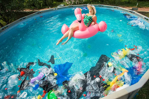 Problema de lixo, reciclagem de plástico, poluição e conceito ambiental - mulher boba nada e se divertir em uma piscina poluída. Garrafas e sacos de plástico flutuam perto dela — Fotografia de Stock