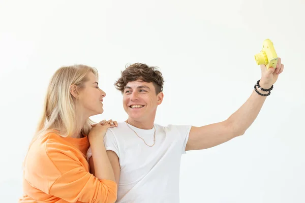 Divertida joven pareja loca rubia y un chico hipster tomando una selfie en una cámara de cine de color amarillo vintage posando sobre un fondo blanco. Concepto de pasatiempo de fotografía . — Foto de Stock