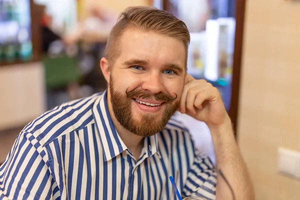 Bonito jovem hipster positivo com um bigode e barba sorrindo posando em um café. O conceito de homens alegres engraçados . — Fotografia de Stock