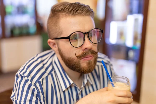 Retrato de un joven guapo con bigote y barba bebiendo café helado en un café sobre un fondo borroso. El concepto de estudiante o los hombres con estilo . — Foto de Stock