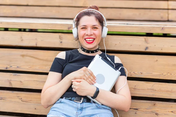 Beautiful young female student is studying using a tablet and headphones while sitting in a park on a wooden bench. Recreation and study concept. — Stock Photo, Image