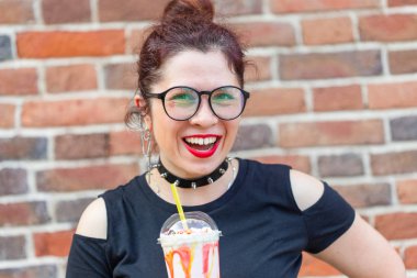 Şık giysiler içinde Sevimli neşeli genç kadın gülüyor ve bir tuğla duvara karşı elinde bir milkshake tutarak. Şehirde bir yaz tatili kavramı.