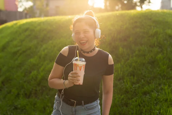 Beautiful sensual young woman listens to music with headphones and holds a milkshake in her hands while walking through the park on a sunny evening. Weekend getaway concept.