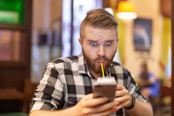 Sorprendido joven hipster bebiendo cacao de una paja y mirando los mensajes en las redes sociales mientras estaba sentado en una mesa en un café. El concepto de chat y redes sociales . — Foto de Stock