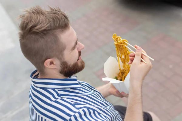 口ひげとあごひげを持つ若い男性のヒップスターは、白い弁当箱から木の箸で中華麺を食べています。アジア料理コンセプト. — ストック写真