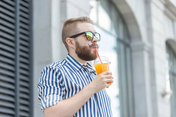 Vista lateral de un joven empresario bastante alegre con un bigote elegante y una barba con jugo en las manos, caminando por la ciudad después de un día de trabajo. Concepto de positivo y descanso . — Foto de Stock