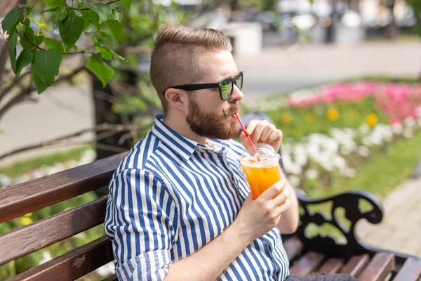 Retrato de un joven positivo y alegre con un vaso de jugo con una pajita mientras camina por el parque en un cálido día soleado de verano. El concepto de descanso después del estudio y el trabajo los fines de semana . — Foto de Stock