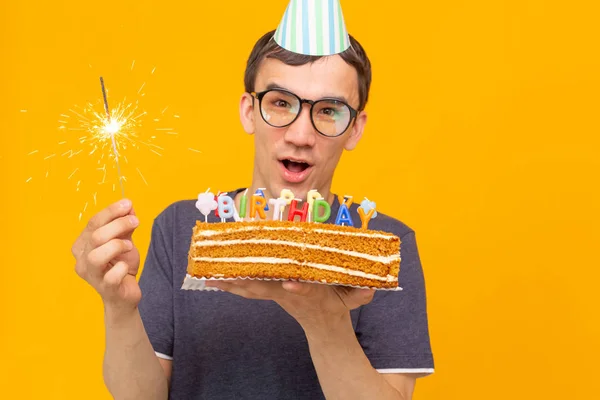 Divertente ragazzo positivo in occhiali tiene tra le mani una torta fatta in casa con l'iscrizione buon compleanno in posa su uno sfondo giallo. Concetto di vacanze e anniversari . — Foto Stock