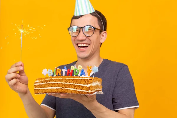 积极有趣的年轻亚洲家伙与帽子和燃烧的蜡烛和自制的蛋糕在他的手中摆出一个黄色的背景。周年纪念和生日概念. — 图库照片