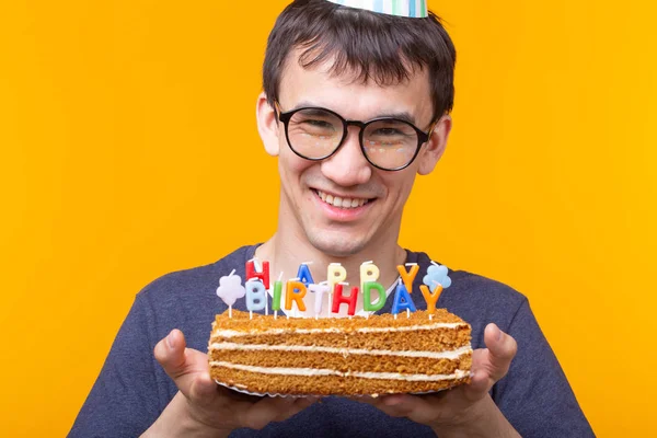 Ritratto di un simpatico ragazzo positivo con un tappo di carta e occhiali che tiene tra le mani una torta fatta in casa di congratulazioni su uno sfondo giallo. Concetto e divertimento e celebrazione . — Foto Stock